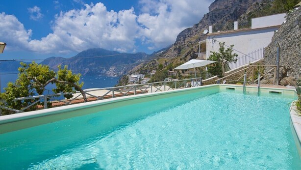Una piscina con agua azul cristalina, rodeada por un edificio blanco con balcón, con montañas al fondo.