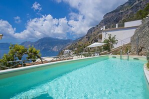 Una piscina con agua azul cristalina, rodeada por un edificio blanco con balcón, con montañas al fondo.