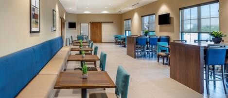 A modern dining area with long tables and blue chairs, a counter with a TV, and large windows.