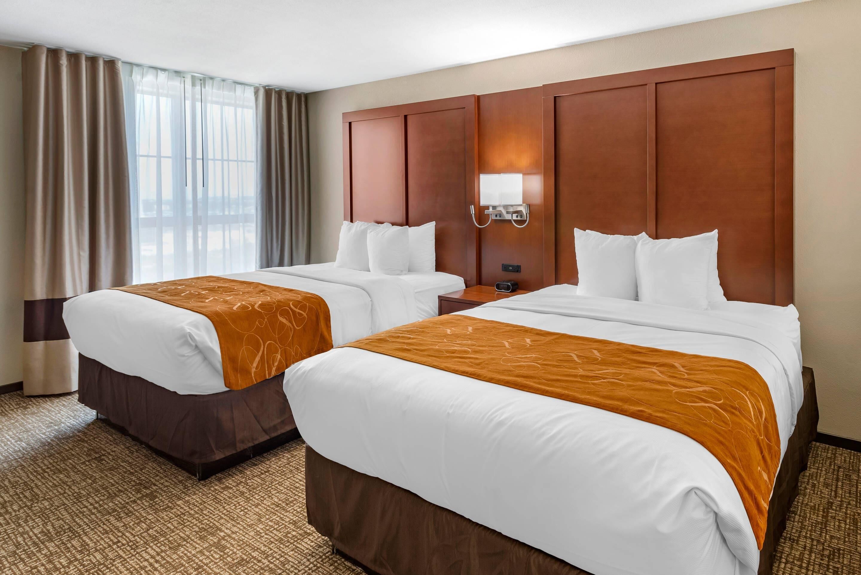 A hotel room with two beds, a large window with curtains, a wall-mounted lamp, and a carpeted floor.