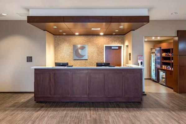 A reception desk with a wooden finish, a stone wall with a logo, and a refrigerator with snacks. - Comfort Suites Midland West (Midland)