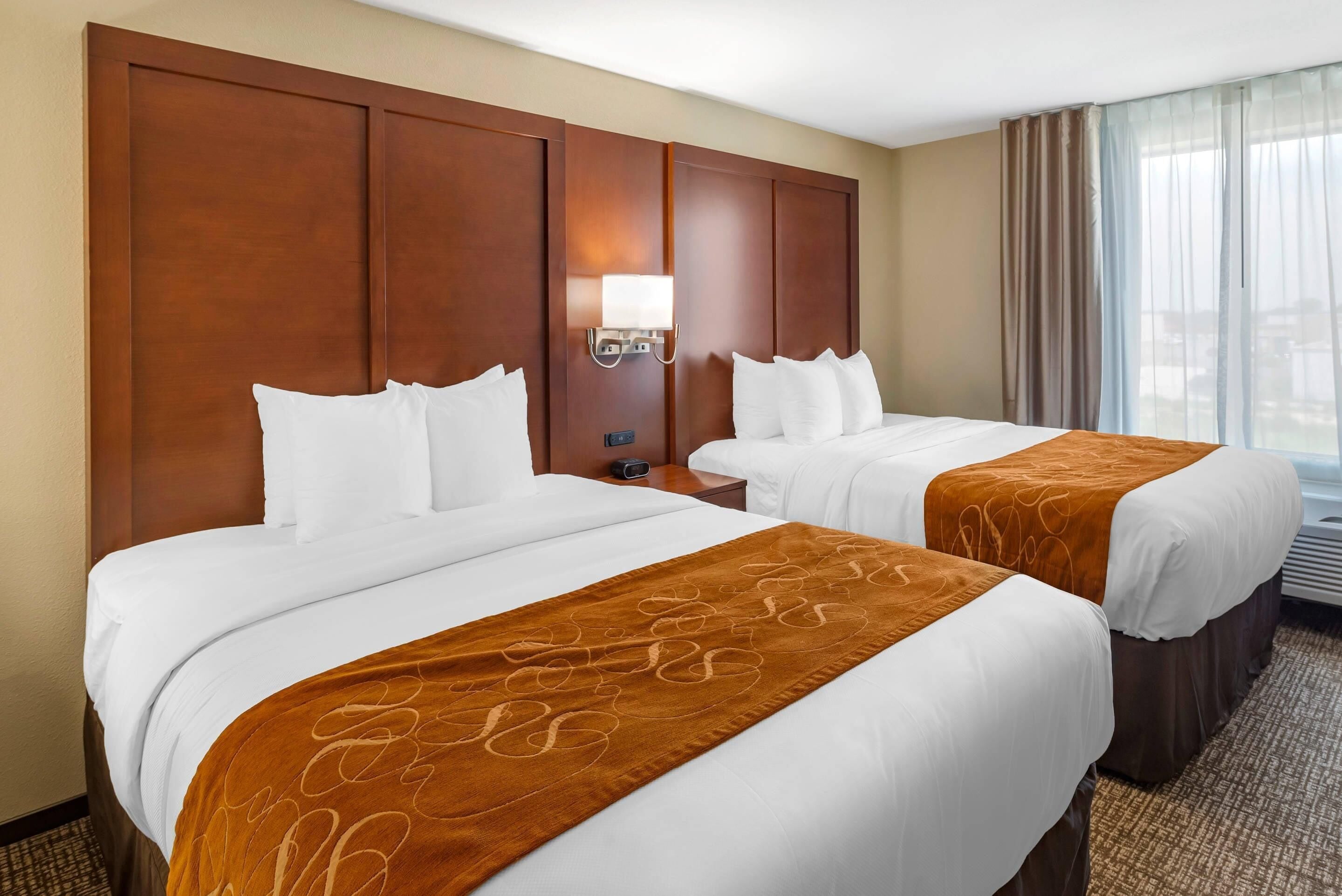 A hotel room with two beds, a nightstand, a lamp, and a window with curtains.