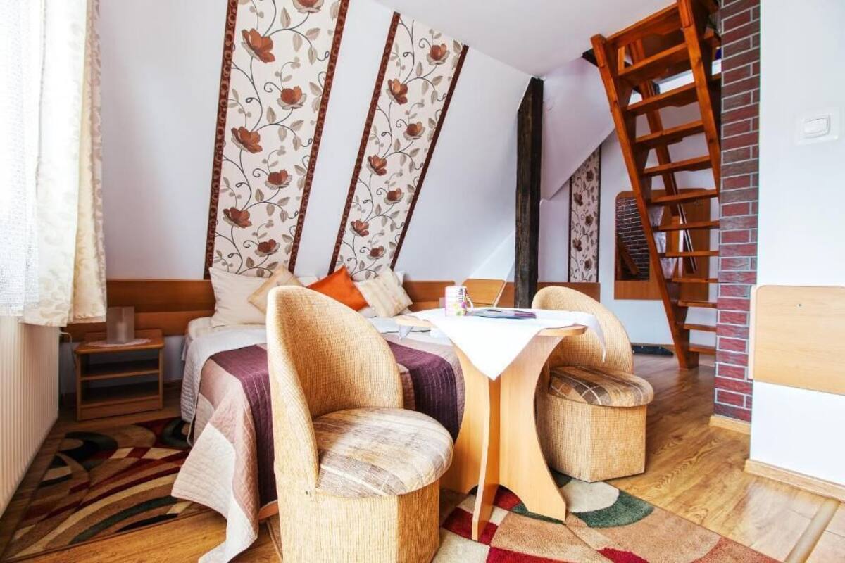 A cozy bedroom with a bed, two chairs, a small table, and a wooden ladder against a brick wall.
