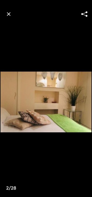 Une chambre à coucher avec un lit, des oreillers, une couvre-lit verte, une table de chevet, un miroir, une étagère avec des objets décoratifs et une plante en pot.