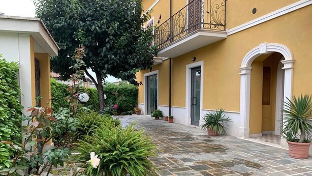 A courtyard with a yellow building, potted plants, and a stone floor. - Victory House 3 (Celano)