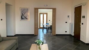 A spacious hotel lobby with a sofa, a coffee table with flowers, a dining area in the background, and a door leading to another room. - Victory House 3 (Celano)