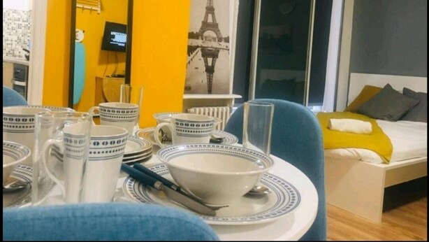 A dining area with a table set for two, a bed with a yellow pillow, and a large window with a view of the Eiffel Tower. - Forset Court (London)