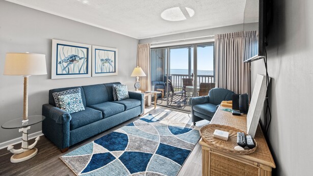 A modern living room with a blue sofa, a coffee table, a TV, and a balcony with curtains.