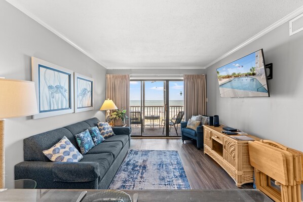 A modern living room with a grey sofa, a blue patterned rug, a wooden console table, and a large window offering a view of the beach.