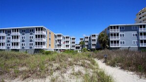 A beachfront residential complex with multiple-story buildings, a sandy path leading to the beach, and a clear blue sky.