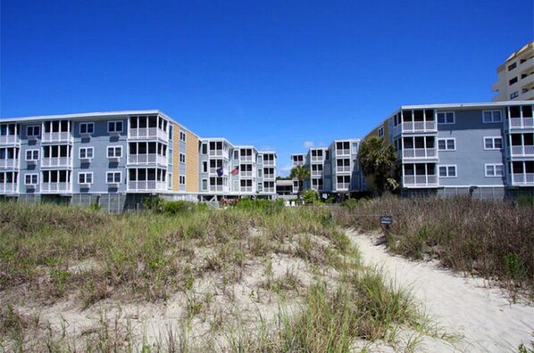 A beachfront residential complex with multiple-story buildings, a sandy path leading to the beach, and a clear blue sky.