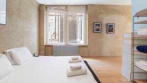 A bedroom with a large bed, a radiator, a window with curtains, and a shelving unit with towels.