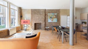A spacious room with a brick fireplace, a dining area with a long table and chairs, a kitchen area with modern appliances, and a large window providing natural light.