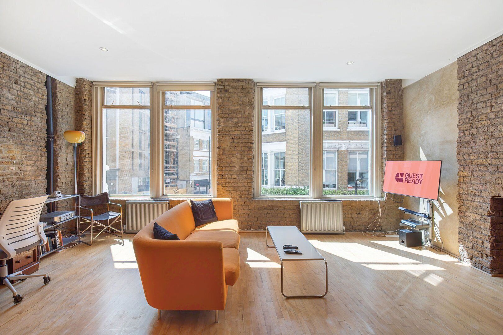 A modern living room with a brick wall, large windows, a sofa, a coffee table, and a TV displaying "GUEST READY".