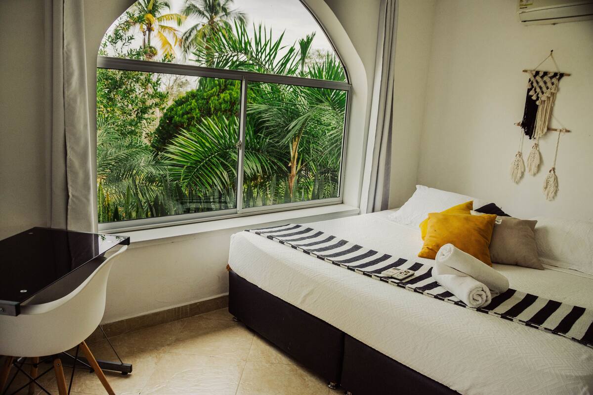 A bedroom with a bed, a desk, and a window with a view of greenery.
