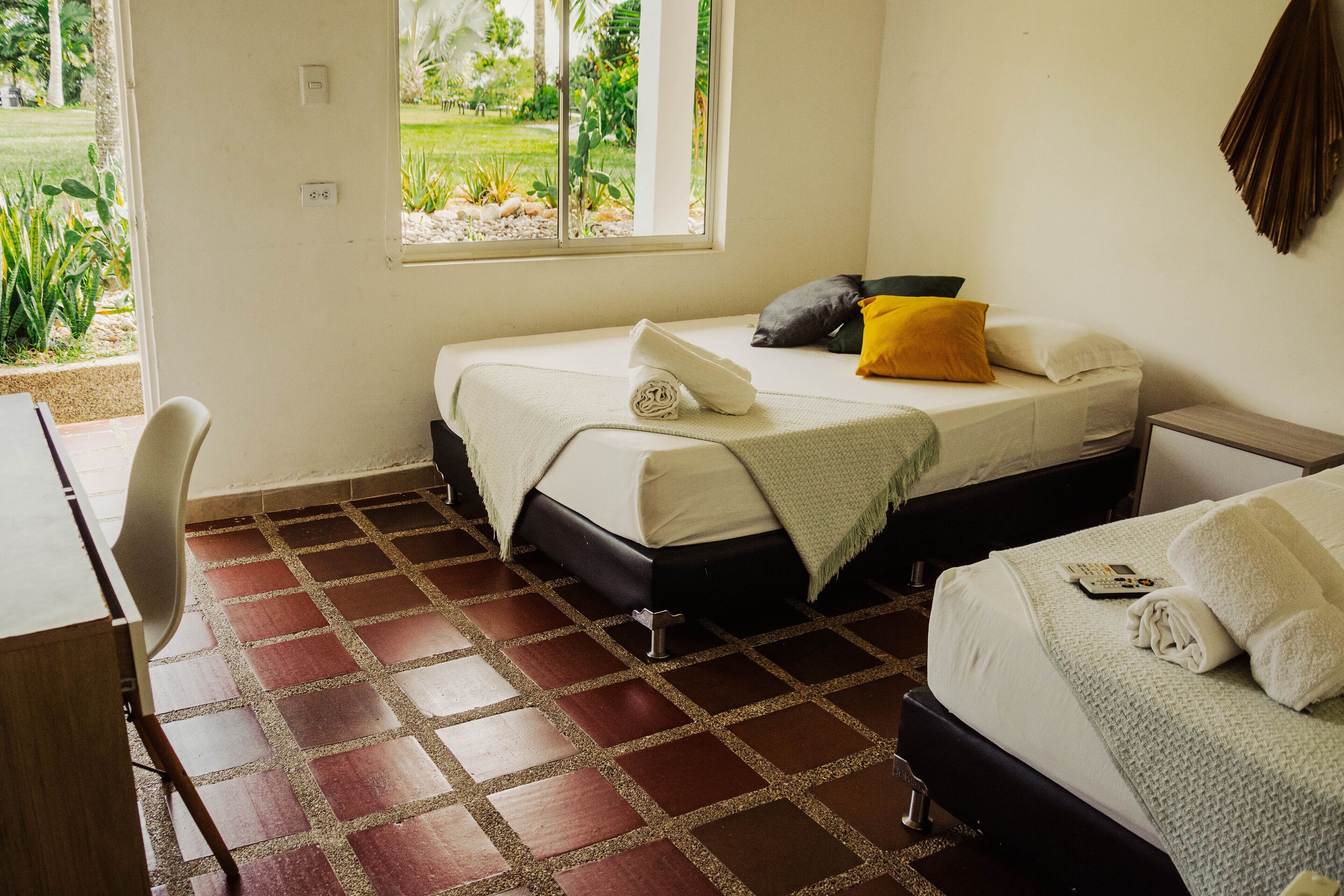 A room with two beds, a desk, and a chair. There is a window with a view of greenery outside.
