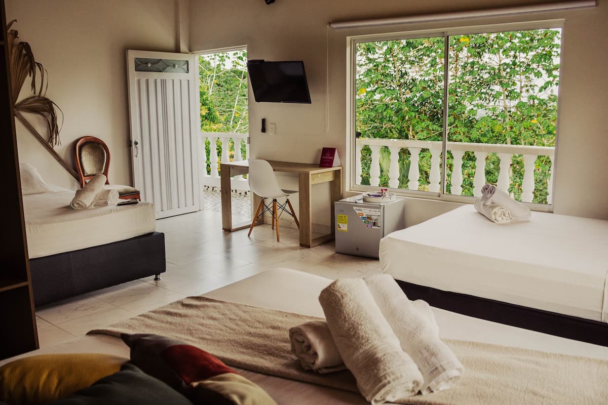 A hotel room with a bed, a desk, a chair, a TV, and a balcony with a view of greenery.