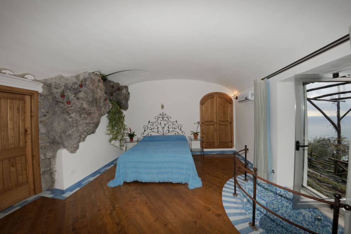 A bedroom with a bed, a wooden door, a stone wall, and a view of the outdoors.