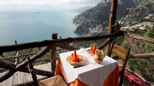 Une table dressée avec des serviettes orange et un vase, donnant sur un paysage côtier avec des bâtiments et des falaises.