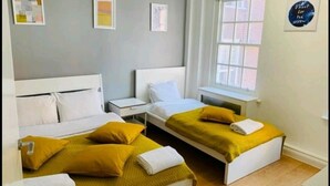 Une chambre d’hôtel avec deux lits, chacun doté d’une tête de lit blanche et d’une couverture jaune. On y trouve deux tables de chevet, une fenêtre avec vue et un mur orné de tableaux.