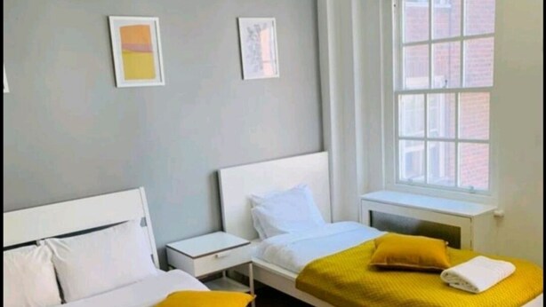 Une chambre d’hôtel avec deux lits simples, chacun doté d’une tête de lit blanche et d’une literie jaune. On y trouve deux tables de chevet, une fenêtre avec des stores et un climatiseur fixé au mur.