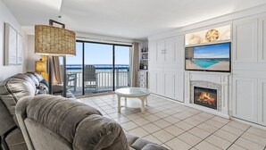 A modern living room with a sofa, a glass coffee table, a fireplace, a TV mounted on the wall, and a balcony with a view of the sea. - Oceans 415-Spacious Oceanfront 1BR- Outdoor Pool (North Myrtle Beach)