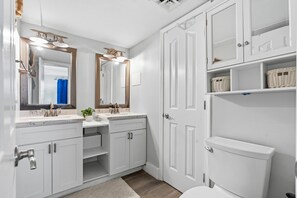 A bathroom with white cabinetry, a double sink, a toilet, and a mirror with lights. - Oceans 415-Spacious Oceanfront 1BR- Outdoor Pool (North Myrtle Beach)