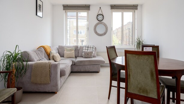 A living room with a grey sectional sofa, a dining table with chairs, and a large window with blinds. - GuestReady - Amazing flat in Trendy Hoxton (London)