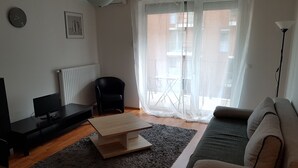 Un soggiorno con un divano, un tavolino, una TV e una poltrona. Si vede una finestra con tende leggere e vista sull'esterno.