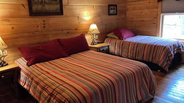 A cabin with two beds, each with striped bedding and a red pillow. There is a lamp on each nightstand, and a framed picture on the wall.