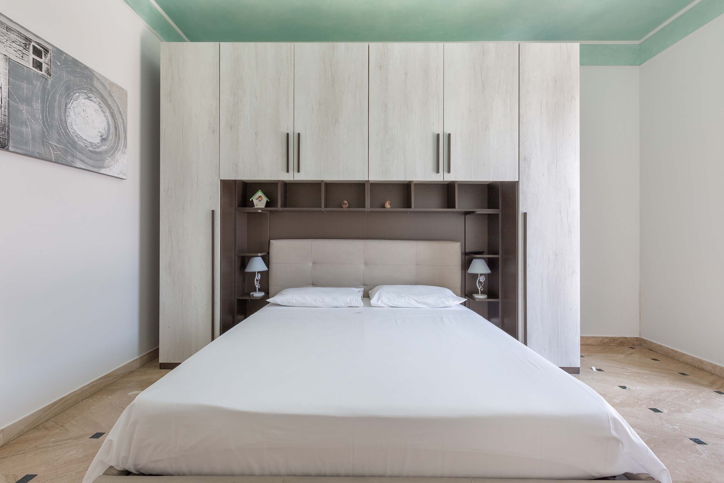 Une chambre à coucher avec un grand lit, une tête de lit dotée de compartiments de rangement et une étagère murale agrémentée d’objets décoratifs.