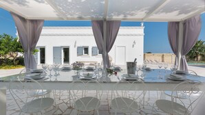 Un espacio de comedor al aire libre con una mesa blanca dispuesta para seis personas, rodeada de sillas blancas, bajo una pérgola con cortinas grises y un edificio blanco al fondo.