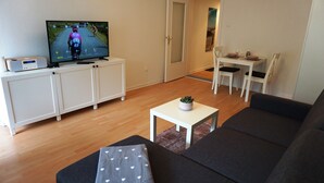 A living room with a sofa, a small table with a plant, a TV displaying a cycling race, and a dining area with chairs and a table. - Central location, close to the train station and the fjord, bus stops nearby (Kiel)