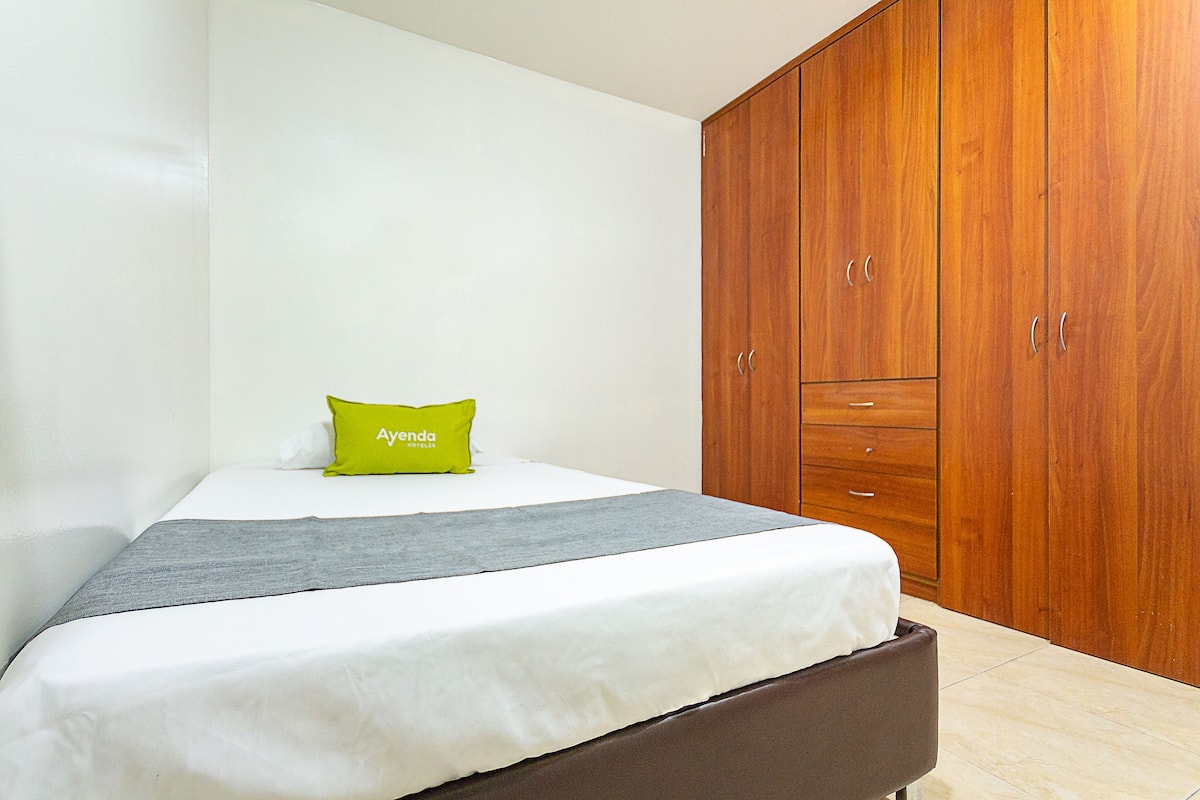 Un dormitorio con una cama, un armario de madera y una almohada verde con la etiqueta 'Ayenda Hotels'.