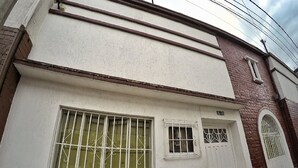 Un bâtiment à la façade blanche, doté d’une grande fenêtre avec des barreaux en métal, d’une fenêtre plus petite également munie de barreaux, et d’une porte.