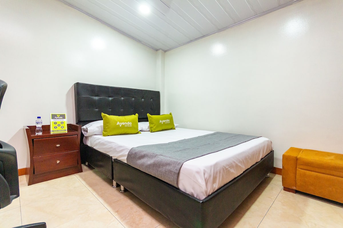 Una habitación de hotel con una cama, una mesita de noche con una botella y un folleto informativo, una silla amarilla y un aparador de madera.