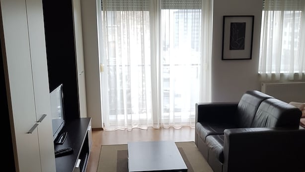 Een woonkamer met een zwarte bank, een salontafel, een tv op een dressoir en een groot raam met dunne gordijnen.