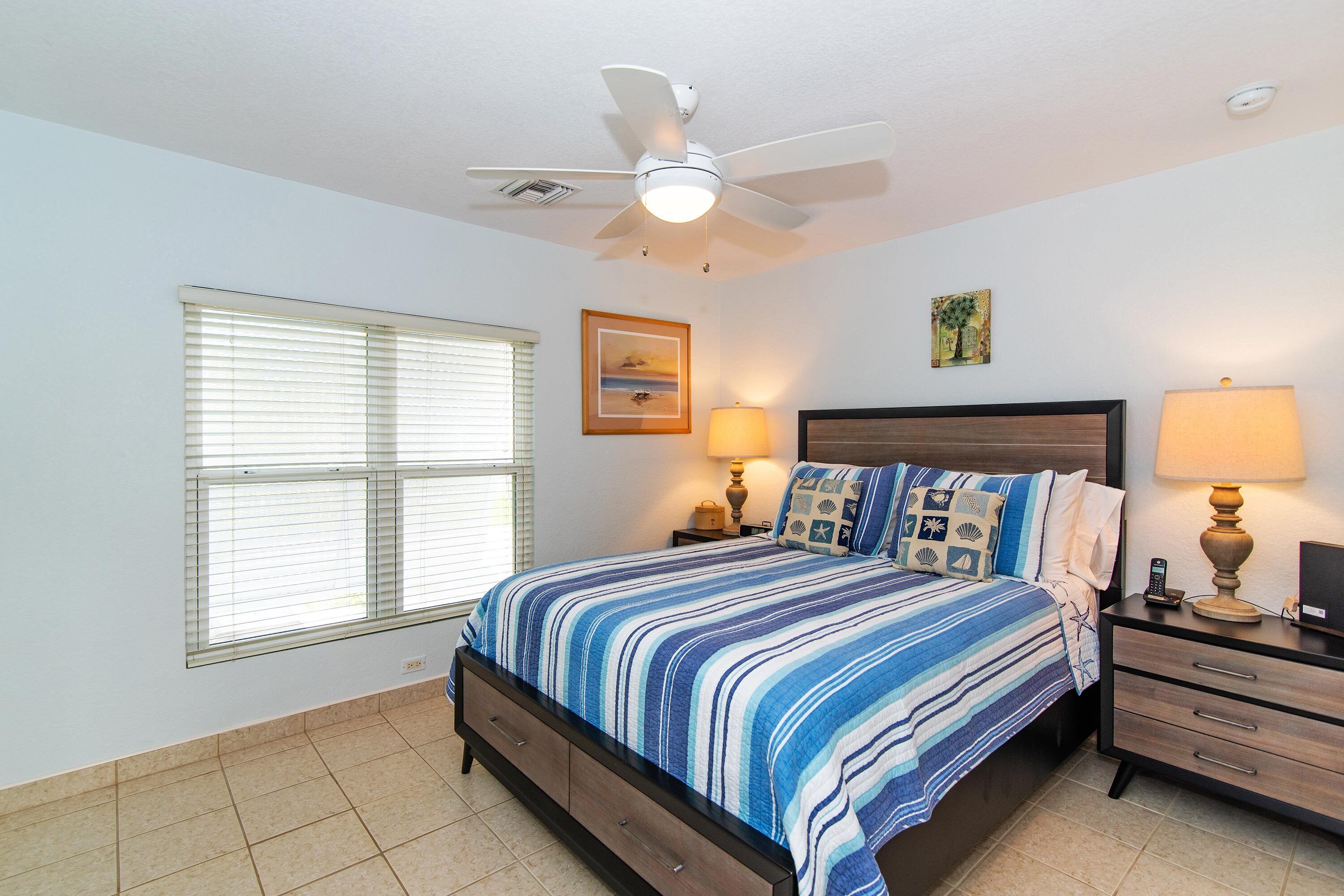 A bedroom with a bed, nightstands, a ceiling fan, a window with blinds, and a wall-mounted picture.