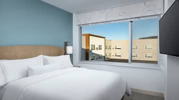 A modern hotel room with a large bed, a window with a view of buildings, and a flat-screen TV mounted on the wall.