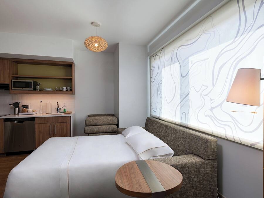 Una habitación de hotel moderna con una cama, una mesita redonda, un sofá, una zona de cocina con microondas y un ventanal grande con cortina estampada.