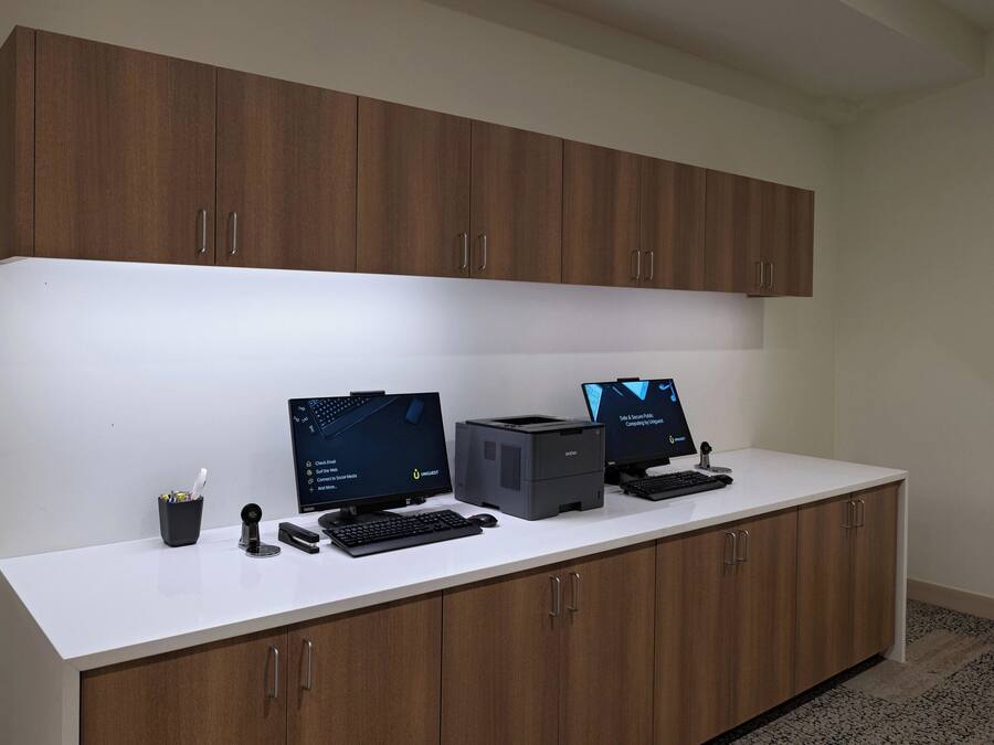 Un espacio de trabajo con dos monitores de ordenador, una impresora y un teclado sobre un escritorio blanco, con armarios de madera arriba.