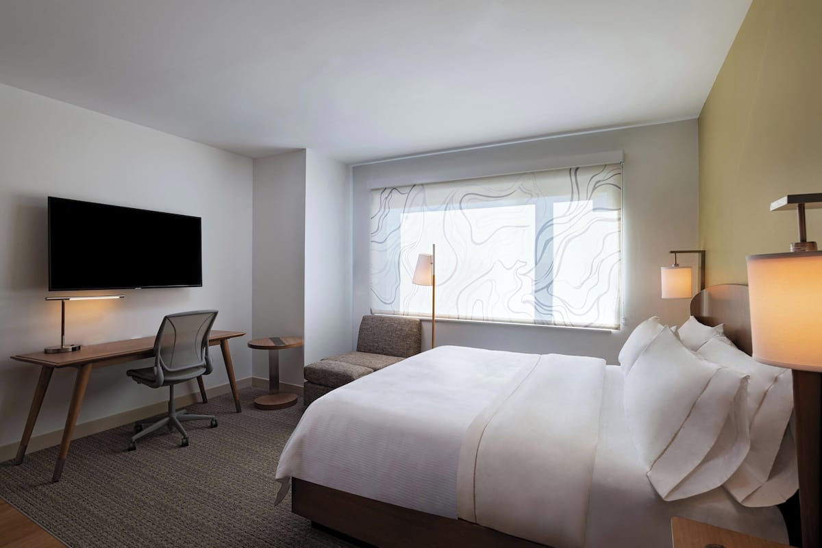 A hotel room with a bed, a desk with a chair, a television, and two lamps.