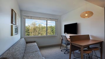 Een moderne woonkamer met een groot raam, een bank, een eettafel met stoelen en een flatscreen televisie aan de muur.