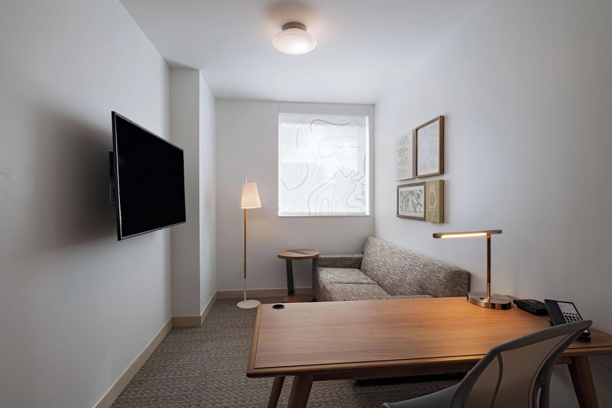 A hotel room with a flat-screen TV mounted on the wall, a wooden desk with a chair, a small round table with a lamp, a sofa, and a window with a view of a building.