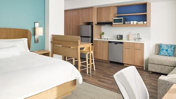A modern hotel room with a bed, a wooden desk, a kitchenette, and a seating area.