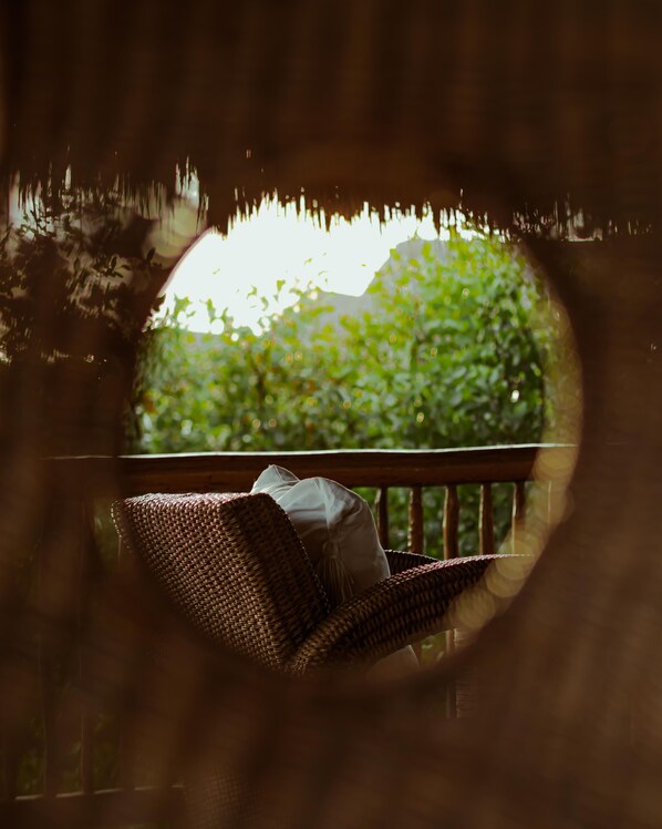 A wicker chair with a cushion on a balcony, framed by a circular window with a view of greenery. - Casa Teka (Tulum)