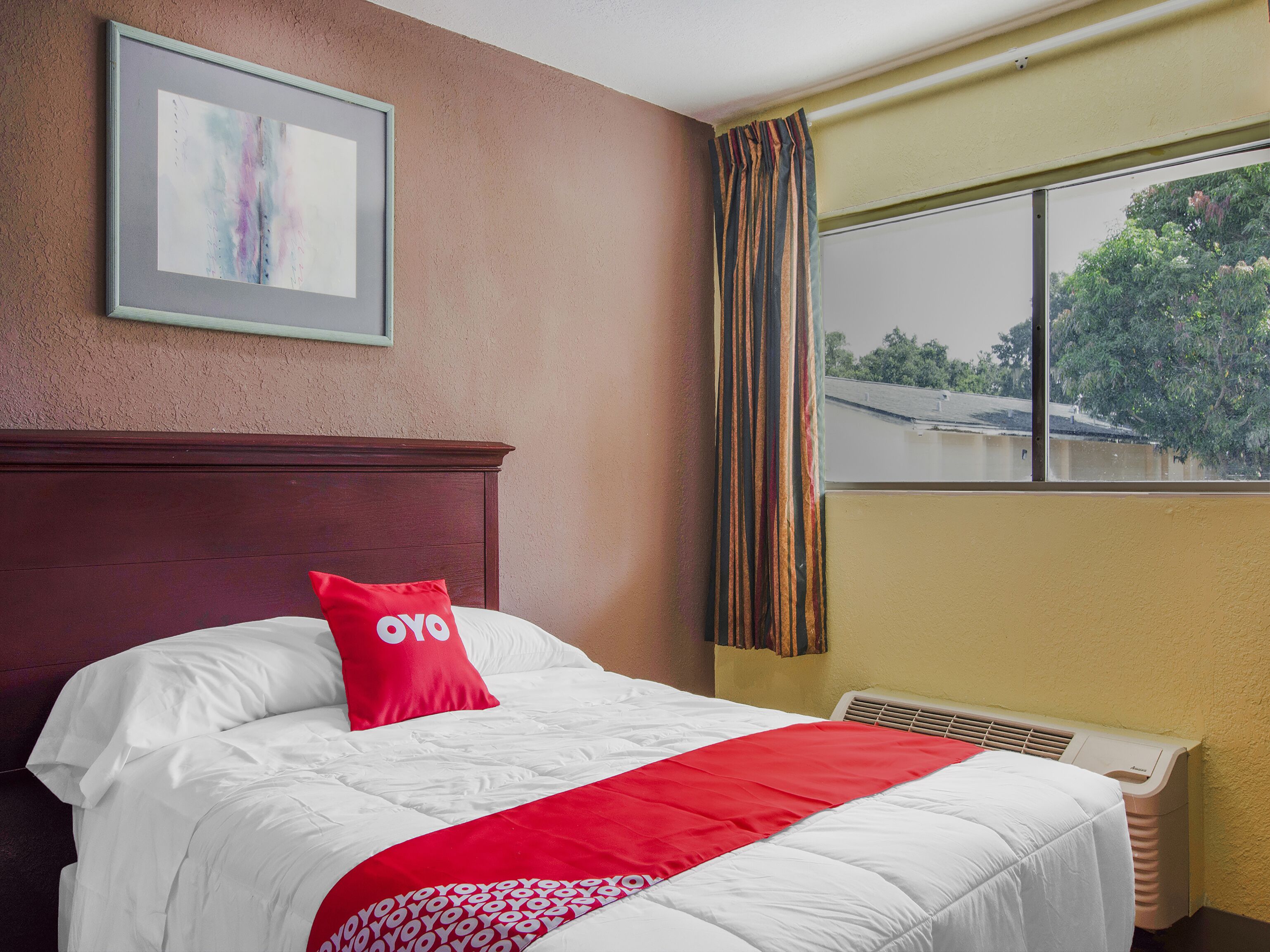 A hotel room with a bed, a framed picture on the wall, a window with curtains, and a radiator.