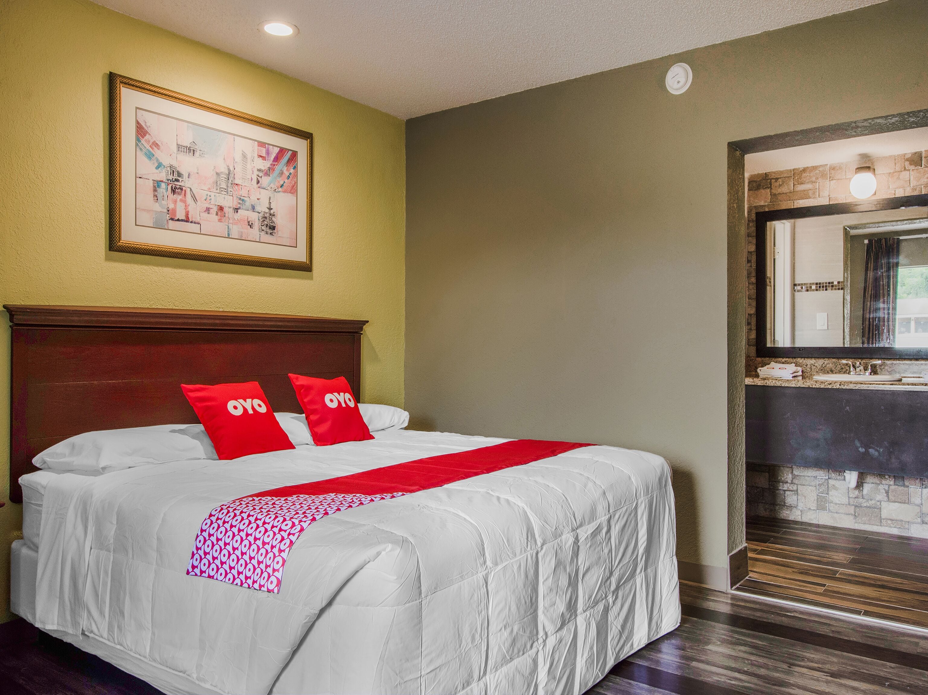 A hotel room with a bed, a painting on the wall, and a bathroom visible through an open door.