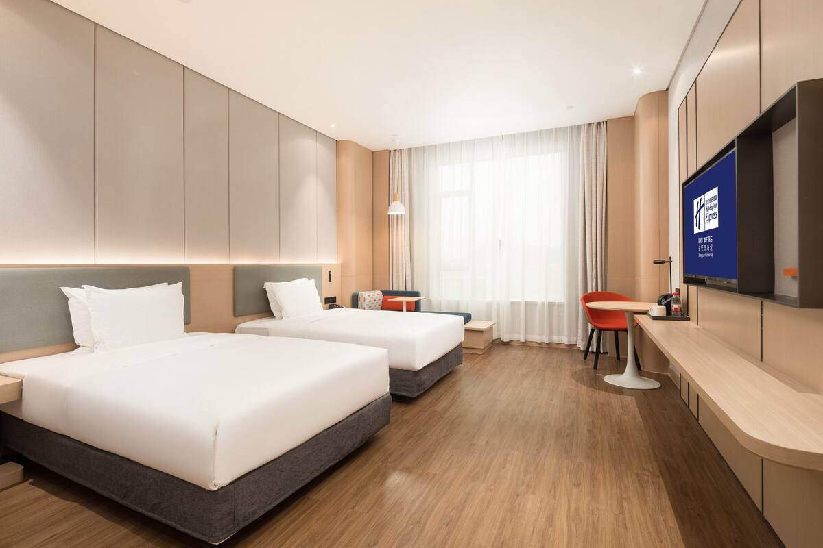 Habitació d'hotel moderna amb dos llits, una taula de menjador petita, una televisió i una finestra gran amb cortines.