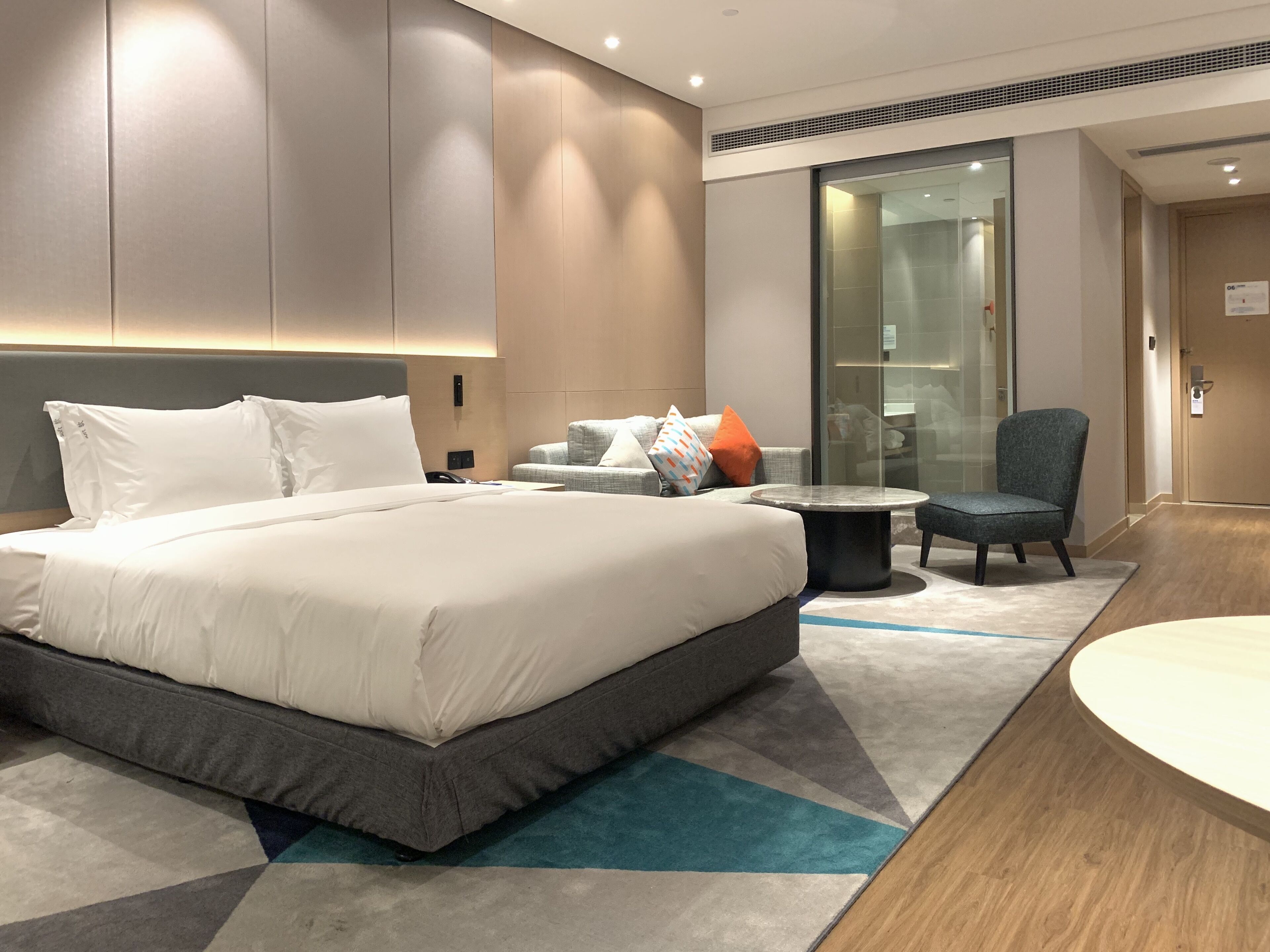 A modern hotel room with a large bed, a seating area with a round table, and a glass partition.